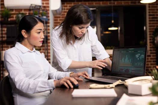 Diverse female colleagues work together at a desk, analyzing financial charts on laptop. Caucasian manager stands and explains details as her asian coworker listens attentively, engaged in discussion.