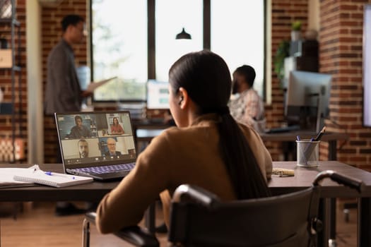 Businesswoman in wheelchair seated at desk with her laptop, having a video conference call with a group of potential investors. Asian female manager conducting a virtual meeting with her remote team.