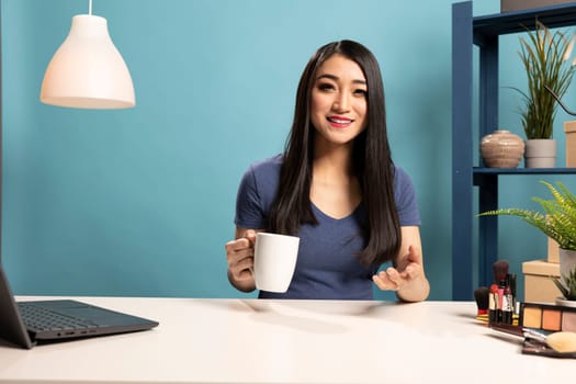 Asian woman smiles and engages with her online audience during a streaming session in blue background studio. The cheerful blogger sits at her desk, holding a coffee cup while looking at the camera.