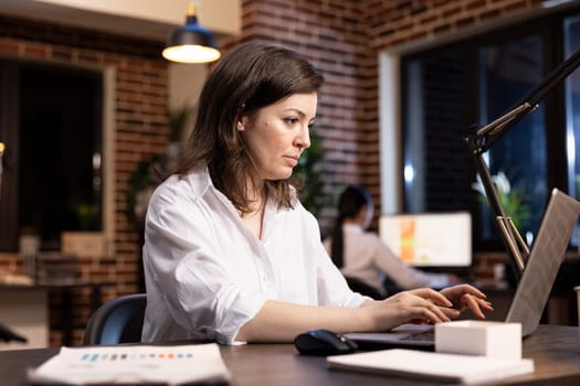 Focused businesswoman works into the night, checking emails and handling responsibilities on her personal computer in office. Caucasian female professional typing on her laptop, planning tasks.