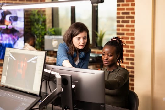African american digital artist and caucasian colleague analyze creative projects on desktop screen in startup company. Multiethnic collaboration at marketing agency, as two women work at office desk.