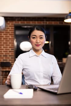 Portrait of asian female executive gripping coffee cup and looking at camera. Professional woman sitting at desk with her laptop and notepad, portraying readiness for late night work in modern office.