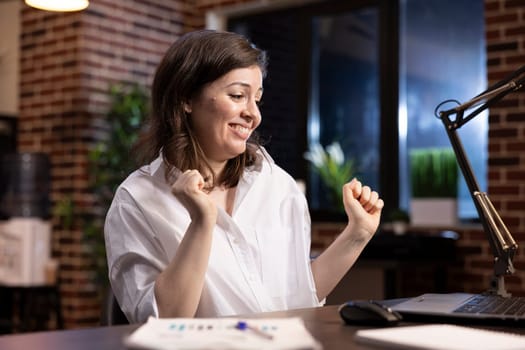 Cheerful businesswoman celebrates at her desk, smiling and dancing on her chair while looking at her laptop. Her energy reflects success, happiness, and motivation in the workplace.