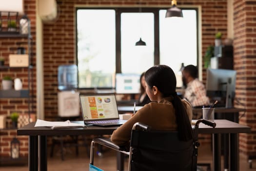 Female employee in a wheelchair working on a laptop, reviewing financial reports and data charts in a startup office. Asian businesswoman using her personal computer in a modern, inclusive workspace.