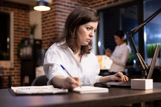 Female professional reviews a project presentation in office, jotting down important notes. Caucasian manager monitors company marketing progress on a laptop, recording key information in a notebook.