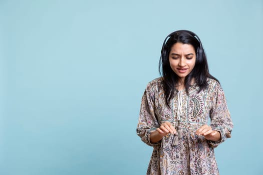 Indian woman enjoying groovy music on headphones and dancing, having fun moving on rhythm against blue background. Pleased young adult does groovy movements wearing audio headset.