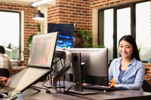 Portrait of asian businesswoman sitting at table and happily working on her computer in modern workspace. Cheerful female professional with her hands on keyboard, typing and looking at camera.