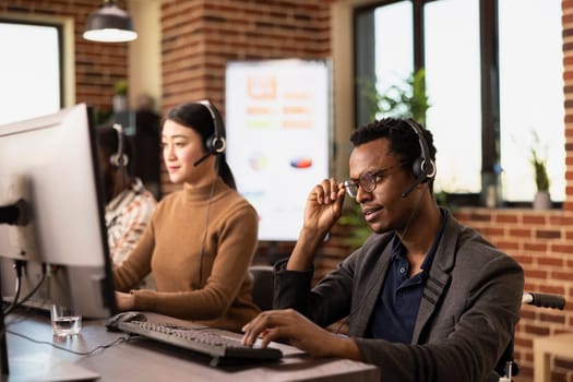 Male wheelchair user touching his glasses and working attentively at his office desk. Black businessman using a headset to communicate with online clients and entering their details on computer.