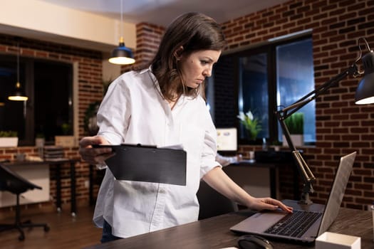 Late at night in a brick wall office, a focused businesswoman stands at her desk, comparing documents on clipboard and laptop. She carefully examines data, ensuring accuracy before making decisions.