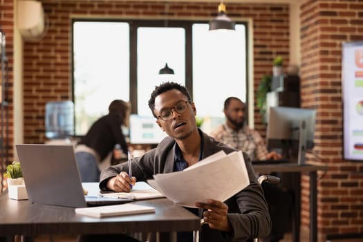 Male wheelchair user with laptop on desk, reviewing financial documents, demonstrating workplace accessibility. Serious black employee with papers and pen in hand, analyzing company performance.