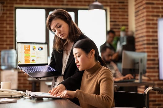 Asian employee at desk participates in business discussions while managing her recovery in modern office. Businesswoman stands with laptop, presenting marketing insights to colleague in wheelchair.