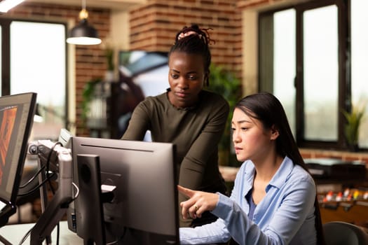 Focused asian and african american coworkers looking at computer, analyzing and discussing on updating graphic designs. Diverse female editors in conversation about digital art in modern office.