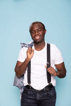 African american man smiles at the camera, standing and holding his denim jacket over his shoulder. Black male model posing against an isolated background, gripping his suspenders.