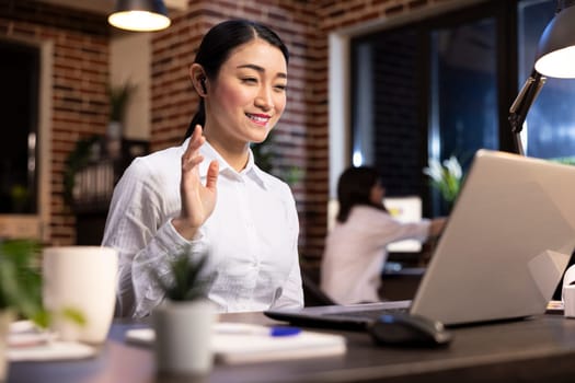 Smiling female manager waving at laptop screen, attending videoconference call with potential client. Asian woman entrepreneur using her personal computer for a virtual meeting with business partners.