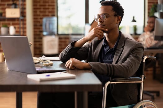 Black businessman in wheelchair holding his chin and concentrating on financial data on his laptop. Male employee with a disability, focused on reviewing a business project plan in startup office.