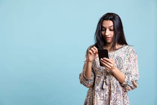 Indian woman happily scrolling on phone to check social media feed, amazed by something. Young relaxed person browsing the internet websites, texting friends against studio background.
