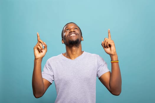 Cheerful african american man pointing upward, looking above as if highlighting a message. Happy male individual in a casual t shirt against a blue background, perfect for an advertisement.