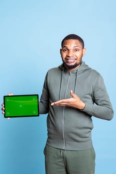 Joyous african american man showing tablet with green screen, doing recommendation against blue background. Upbeat guy pointing at the device with isolated mockup, giving positive feedback.