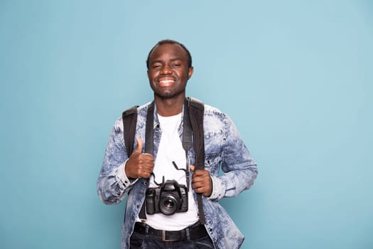 Cheerful black photographer enthusiastically gestures a thumbs up, ready for his next project. African american cameraman smiles brightly, equipped with a backpack and DSLR camera.