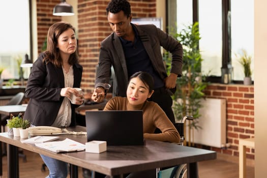 Female accountant in wheelchair analyzing business analytics with colleagues in brick wall office. Multiracial startup team surrounds a laptop on desk, collaborating to improve financial strategies.