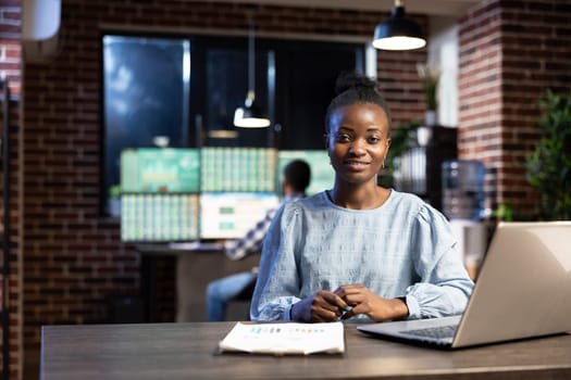 Black woman working late at night in modern office, using her laptop and clipboard at her desk. African american female stock analyst sitting at table and looking at camera, ready for her work shift.