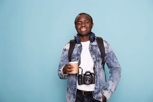 Portrait of male photographer with DSLR and headphones around his neck, confidently posing with one hand in pocket. Black man in casual clothing standing with a cup of coffee and looking at camera.