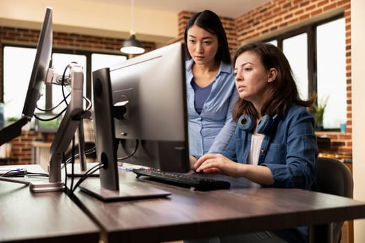 Creative agency female professionals collaborate in production department, brainstorming innovative ideas. Asian developer and caucasian colleague looking at computer screen, reviewing their designs.