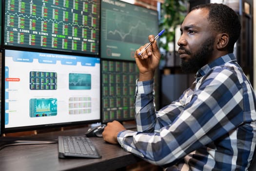 Focused black man working late at a desk, analyzing stock market data on multiple monitors in a modern office. Stockbroker holding a pen as he contemplates and reviews trading statistics.