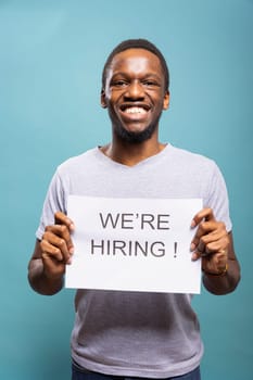 African American businessman holding a job hire sign and standing against a blue background with excitement. Cheerful male individual presenting open recruitment opportunities for candidates.