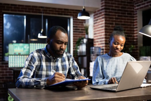 African american financial analyst and stock trader engage in discussion, exchanging insights on company shares. Male and female coworkers review key figures on laptop and clipboard in nighttime office