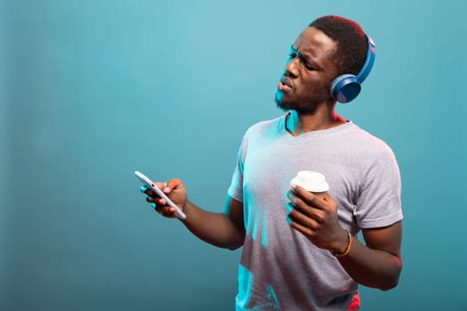 Man wearing blue headphones, holding his coffee and checking his phone with a shocked expression. African american guy with disposable cup reacting in surprise to startling news on his mobile device.