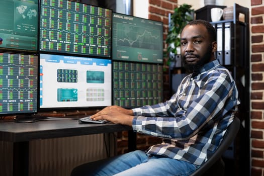 Portrait of black male stockbroker sitting at his desk with computer keyboard and multi screens, ready for a trading session. African american entrepreneur in brick wall office, looking at camera.