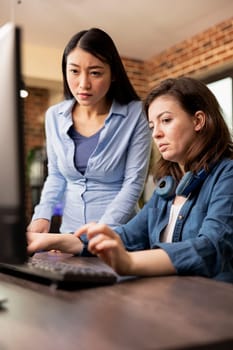 Two diverse women in modern office engaged in a creative discussion, one typing on the keyboard as the other reviews the work on the screen. Asian and caucasian colleagues collaborating on a project.