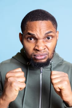Confident powerful young man preparing to fight and box with his fists clenched against blue background. Active muscular male boxer warms up for combat practice and competition, action.