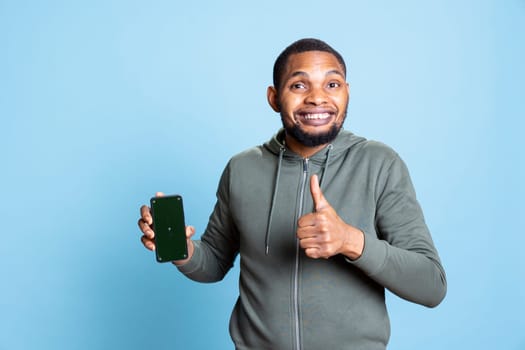Portrait of african american guy showing chroma key screen on smartphone, creating a commercial ad against blue background. Young person smiling and recommending something in studio.