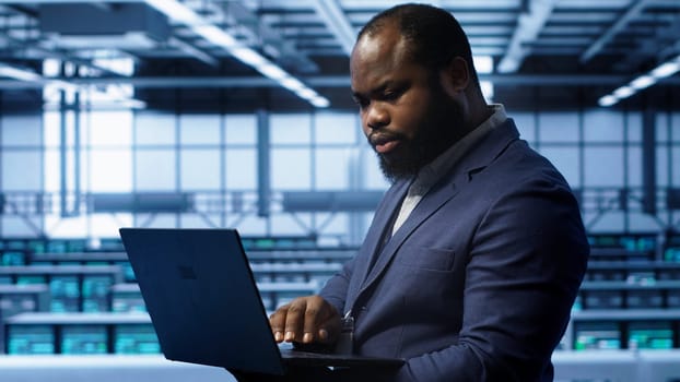 Software developer in server hub checking recovery plan on laptop, monitoring hardware energy consumption. African american worker uses notebook in data center, ensuring rigs are functioning, camera A