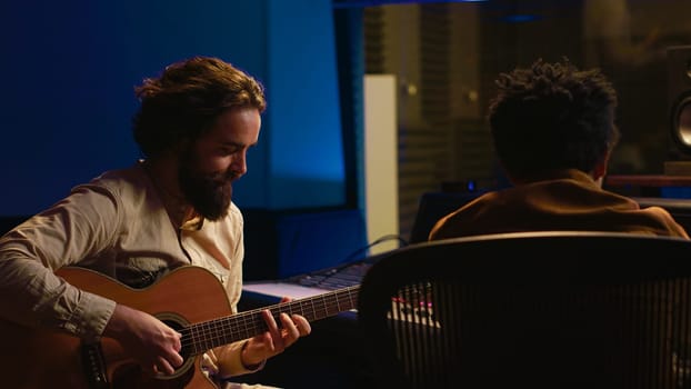 Performer songwriter singing new tracks on his electro acoustic guitar and recording new music in post production. Musician working in collaboration with audio engineer in studio. Camera A.
