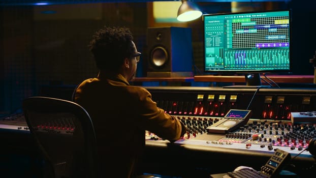 African american sound designer operates control desk mixer and audio software for editing tracks. Producer works with switchers, buttons, sliders, motorized Faders to play hit song. Camera A.