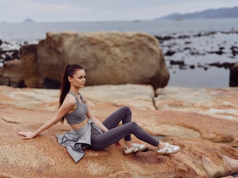 A confident young woman in fashionable activewear sits on a rocky beach, capturing the essence of fitness, relaxation, and natural beauty against a serene waterfront backdrop
