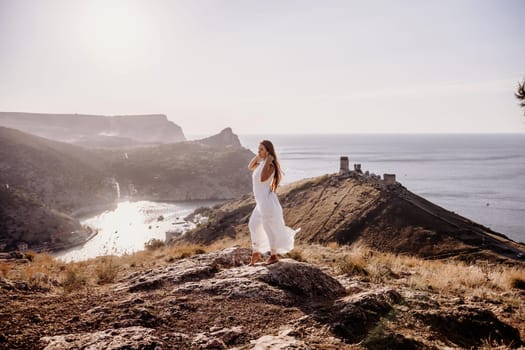 woman stands on a rocky hill overlooking a body of water. She is wearing a white dress and she is in a state of joy or celebration. Concept of freedom and happiness