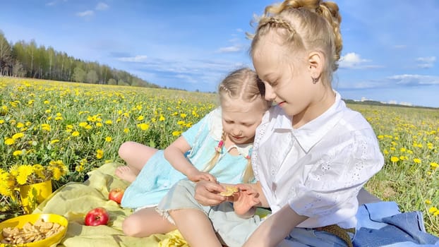 Two siblings happily share snacks during a sunny picnic in a vibrant dandelion field, surrounded by a clear blue sky.