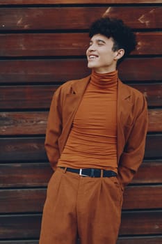 A fashionable young man wearing a rust colored turtleneck and matching suit, smiling in front of a rustic wooden wall