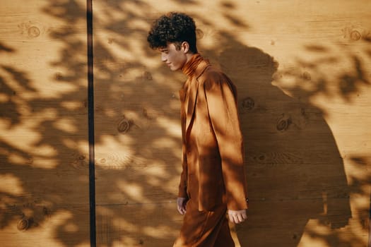A fashionable male model in a brown turtleneck outfit walks gracefully along a wooden wall, with soft shadows creating a tranquil atmosphere