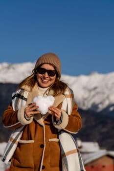 Smiling woman in a brown coat and sunglasses holds a small snow heart against a snowy mountain backdrop on a sunny day.