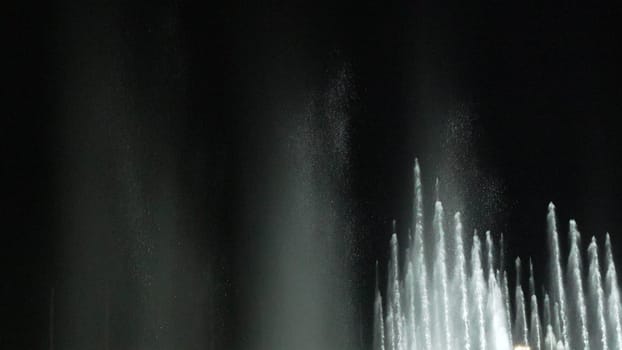 Nighttime view of numerous water fountains shooting high vertical streams of water against a completely dark background. The water appears white and luminous.