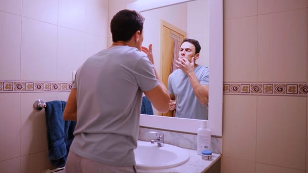 Young man moisturizing his lips with cocoa butter in front of a bathroom mirror
