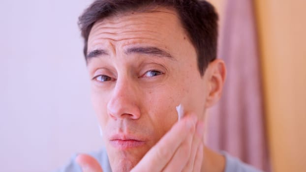 Young man applying moisturizing cream on face for skincare routine