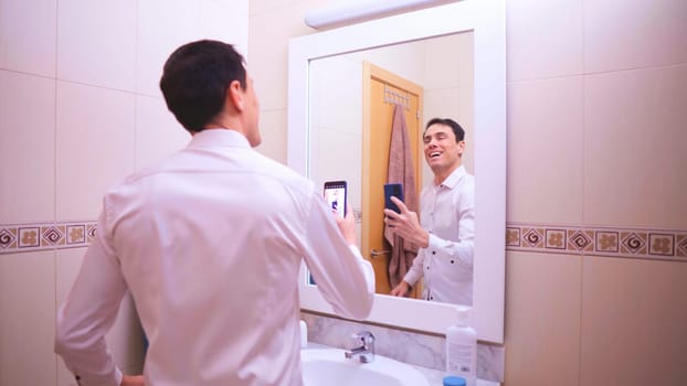 Smiling businessman taking a selfie in the bathroom mirror, capturing his reflection in a white shirt