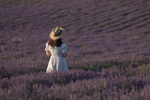 Here's a caption that meets the requirements: Woman in white dress and straw hat sits amidst a vast lavender field at sunset. Back view. Serene, idyllic scene.