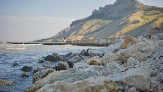 Waves gently washing over rocks on the coast of crimea, with blurred people enjoying the seaside and a mountain in the background